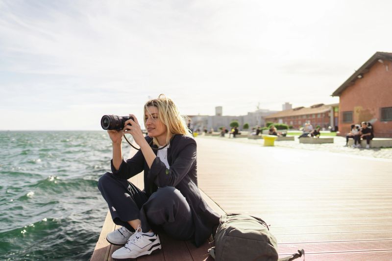 Photo of a young woman, traveling alone and having fun while getting to know the new city. She is sitting on a pier by the sea of a city she is visiting and enjoying the warm springtime weather. Photographing.