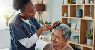 Caregiver gently combs elderly man's gray hair at home, both smiling.