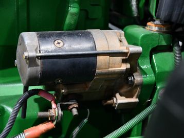 Close-up of an engine starter motor with cables attached and green machinery background.