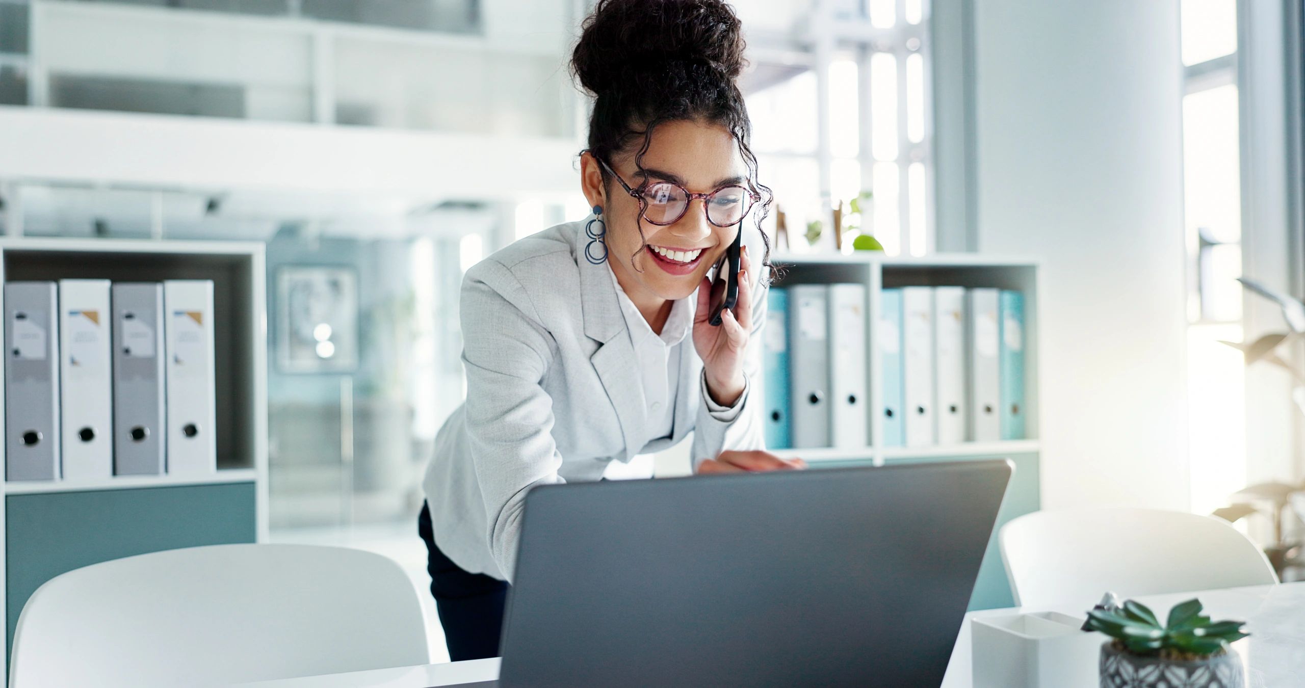 Woman in glasses happily working on laptop while talking on phone in office.