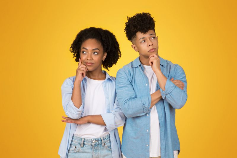 Two pensive thoughtful African American siblings in casual denim attire stand side by side, lost in thought with curious expressions, think, against a bright yellow background