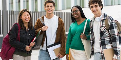 Four diverse students smiling outside a modern building with books and backpacks.