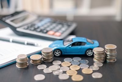 Stacks of coins around a blue toy car with a calculator and pen in the background.