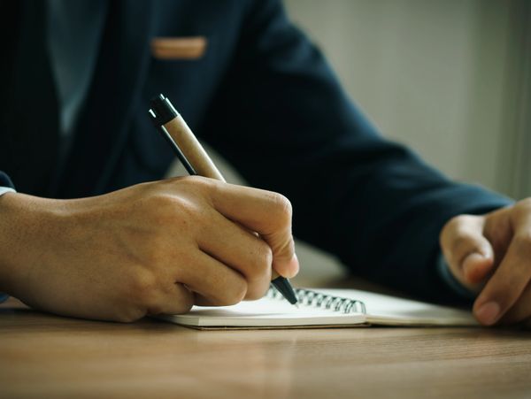 Close-up of a person in a suit writing with a pen in a small notebook.