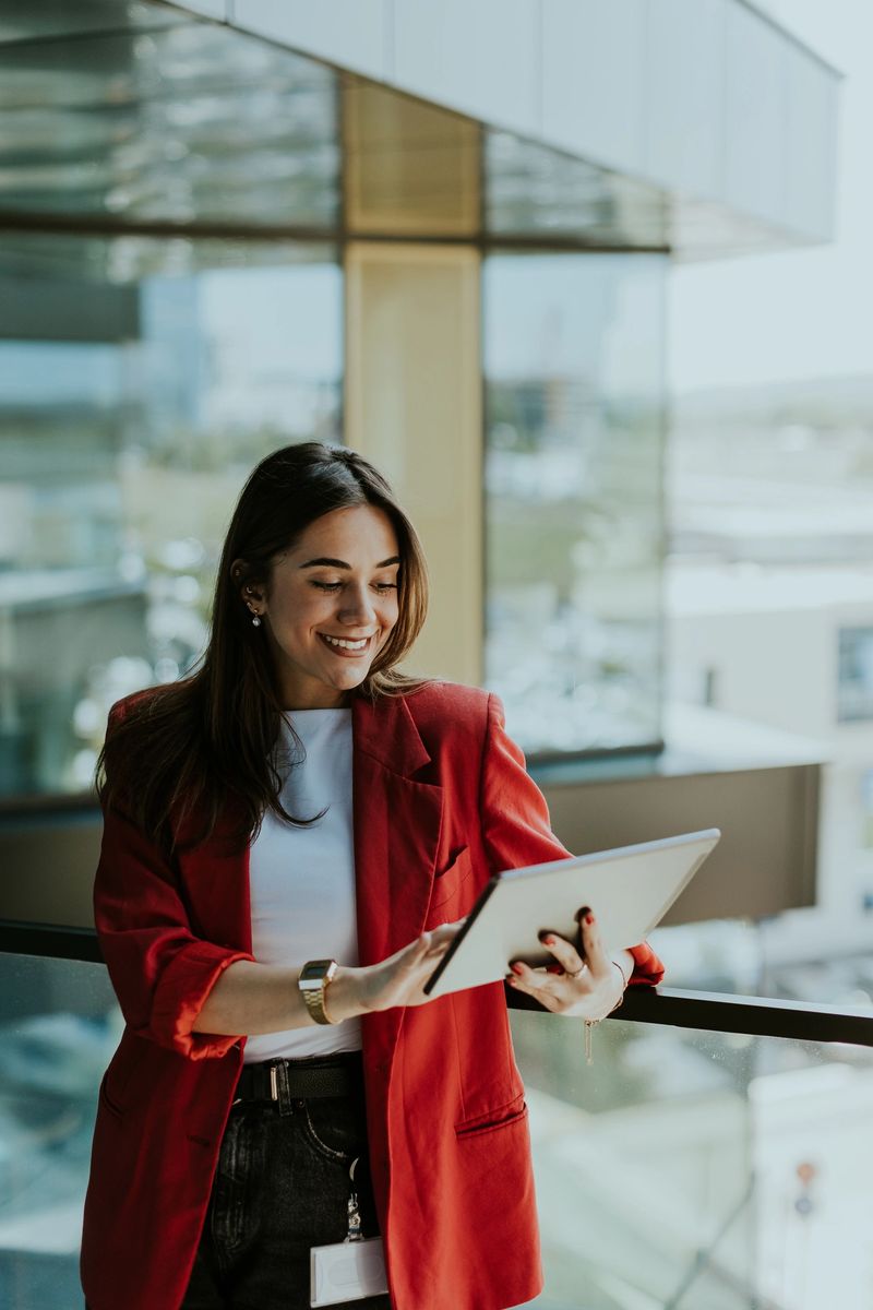 Joyful businesswoman, dressed in a red blazer, interacts with a tablet near a large office window offering a clear view of the city on a sunny day