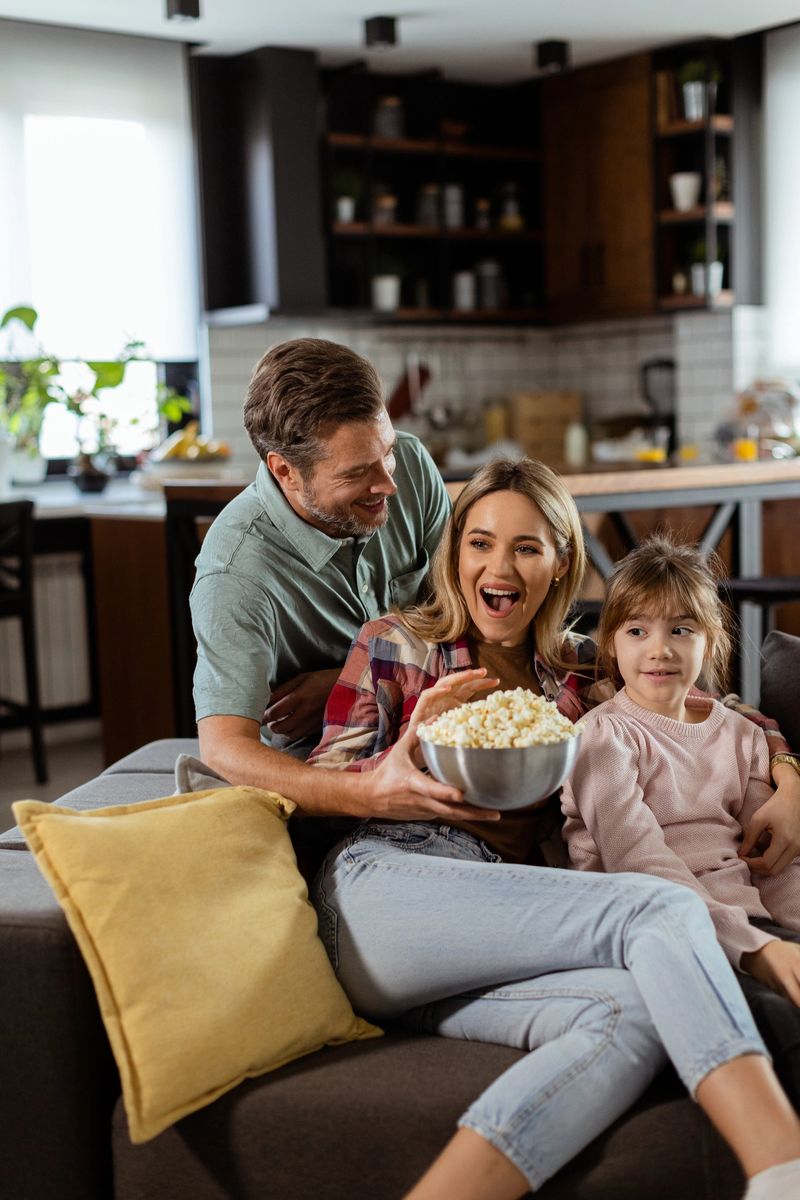 A family of three is comfortably nestled on a couch, their faces reflecting excitement and attentiveness as they share a bowl of popcorn during a suspenseful movie night