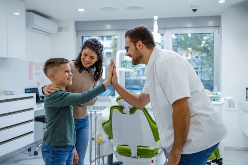 A pediatric dentist celebrates with a young boy, likely after a successful dental checkup, as his mother watches and smiles in the dental office