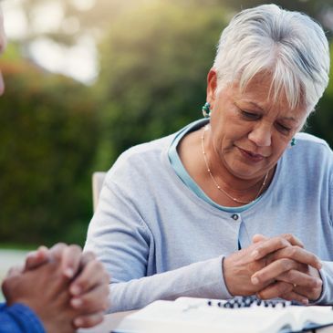 Two elderly people praying together outdoors with a Bible.