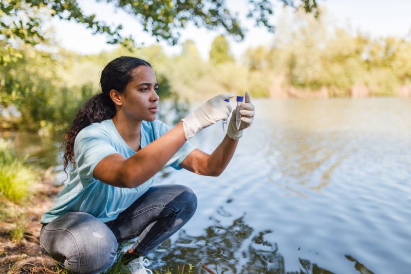 A young woman wearing gloves is collecting water samples from a river to analyze for environmental conservation purposes.