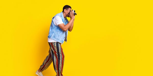 Man in colorful pants taking a photo with a vintage camera against a yellow background.