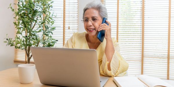 Elderly woman using laptop and talking on phone in a bright room.