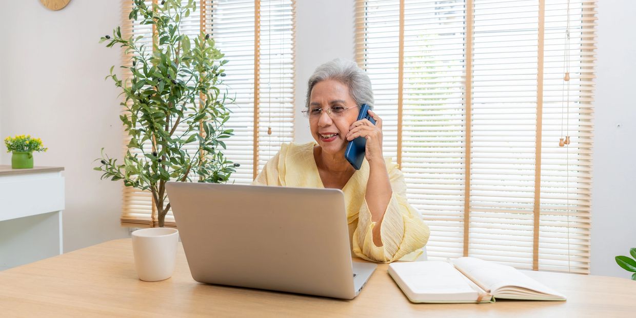 Elderly woman using laptop and talking on phone in a bright room.