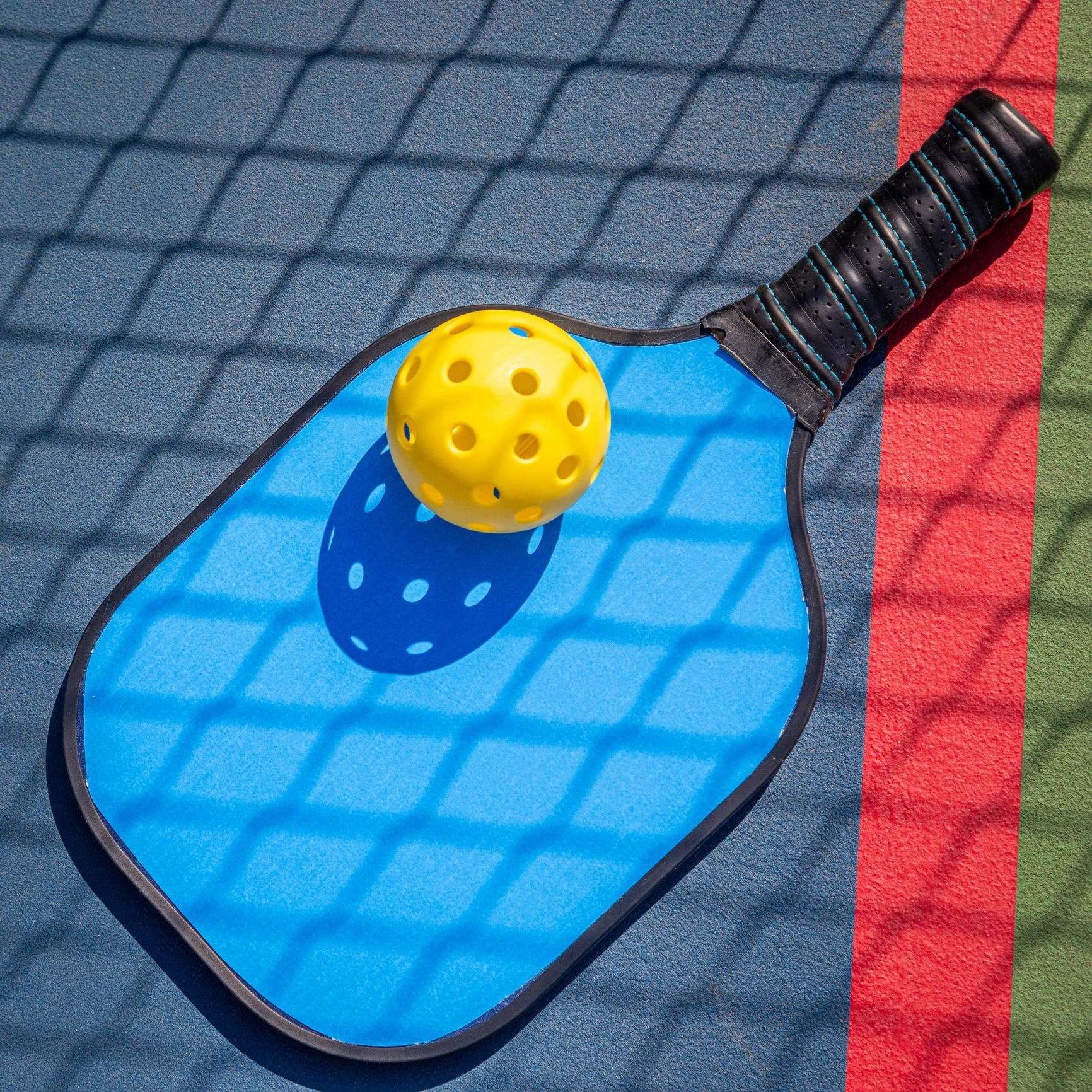 Pickleball paddle and yellow ball on colorful court with net shadow.