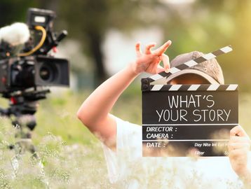 Person holding a clapperboard reading 'What's Your Story' near a film camera outdoors.