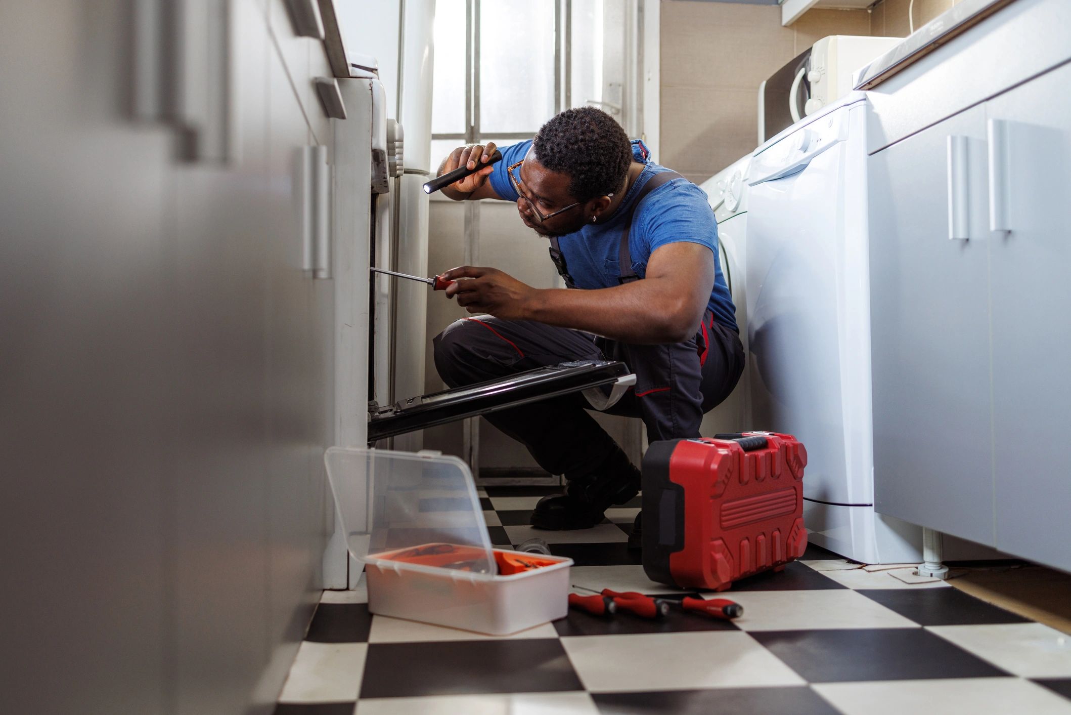 A man repairs an oven with a flashlight and screwdriver in a kitchen.