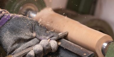 A gloved hand shaping wood on a lathe, creating wood shavings.
