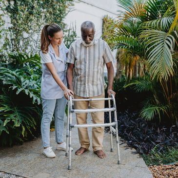 A caregiver assists an elderly man using a walker in a garden.