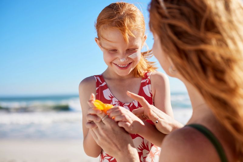 Young mother applying protective sunscreen on daughter nose at beach with copy space. Woman hand putting sun lotion on female child face at sea. Cute little girl with red hair and freckles applying sunblock cream on nose and cheek at seaside.