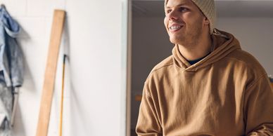 Young man in a beanie and brown hoodie smiling indoors.