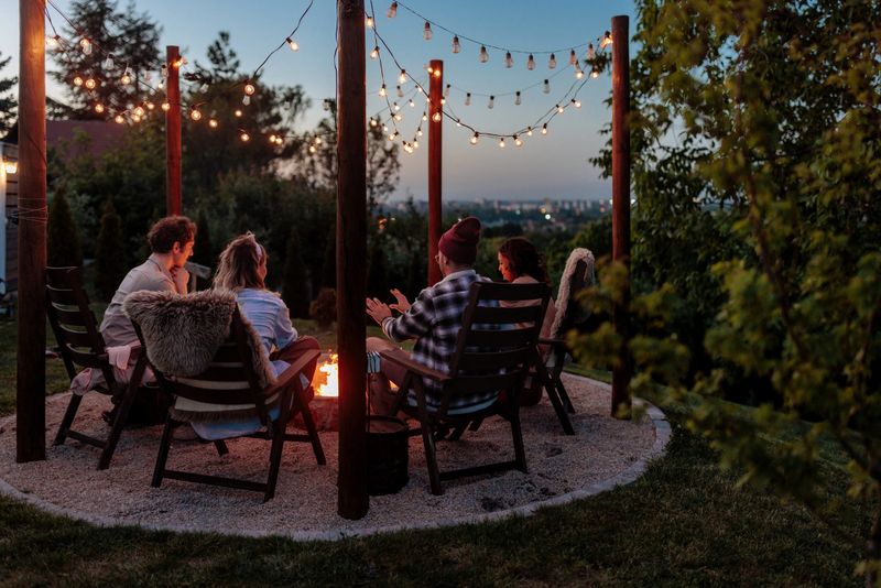 Rear view of a group of four people sitting next to a burning fire pit at sunset in a lush, green garden.