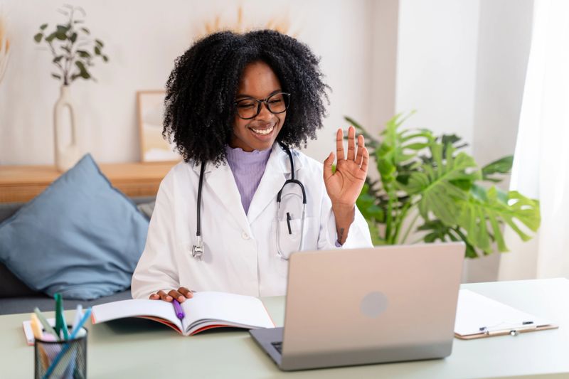 Smiling african american female doctor in white lab coat waving at laptop, conducting online medical consultation from a modern office