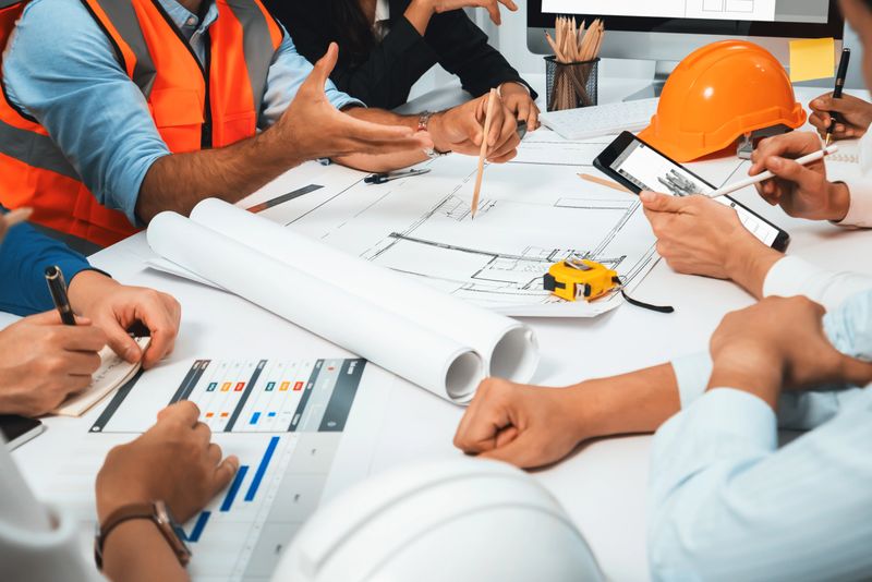 Diverse group of civil engineer and client working together on architectural project, reviewing construction plan and building blueprint at meeting table. Prudent