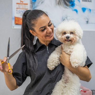 A groomer holding scissors with a fluffy white dog in her arms.