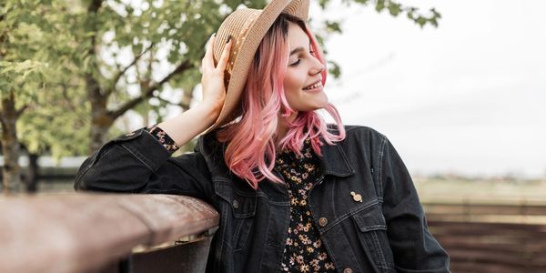 Young woman with pink hair smiling and holding a hat outdoors.