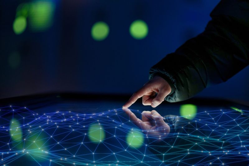 A close-up of a hand touching a digital network interface with blue interconnected nodes and lines, set against a dark background with blurred green lights.