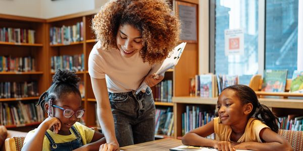 A teacher helps two young girls with reading in a library.