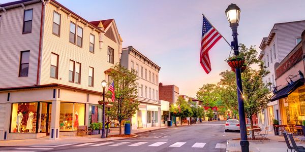 Quiet small town street with American flags and lit storefronts at dusk.