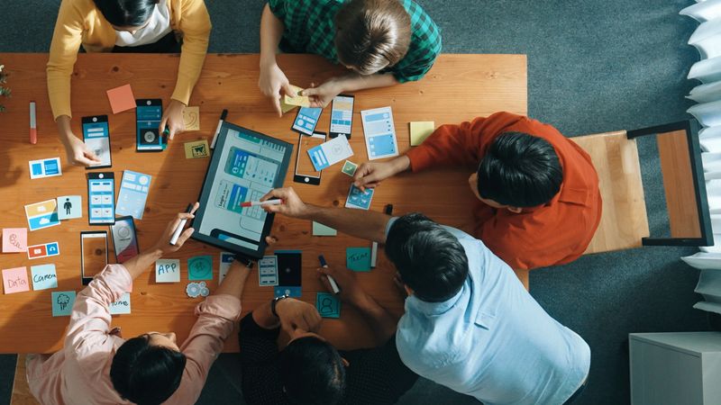 Top view of software developer team looking Ux Ui design for mobile phone interface from tablet while comparing and planning wireframe prototype designing together at creative workplace. Convocation.