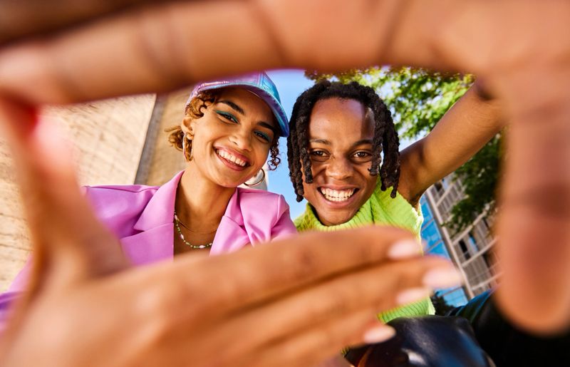 Two Gen Z adults smiling and looking through a hand frame gesture outdoors in an urban environment. Low angle shot.