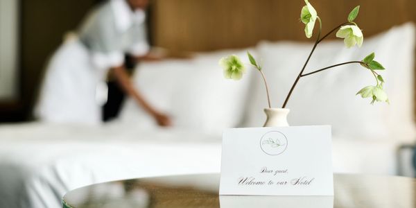 A hotel welcome card with flowers on a table and a maid making the bed.