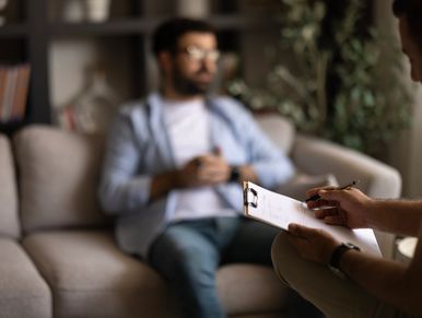 Therapist taking notes while a man talks on a couch.