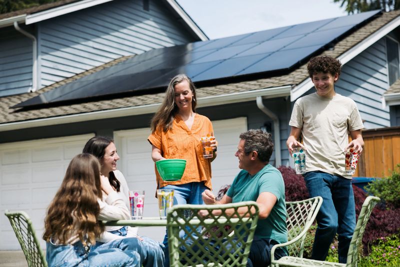 A Caucasian family with three children have fun connecting with one another outside on their lawn, sitting at an outdoor table.  Their solar panel array is visible on the roof of their home, providing green natural energy for the environmentally conscious family.  Sustainable modern lifestyle in action.