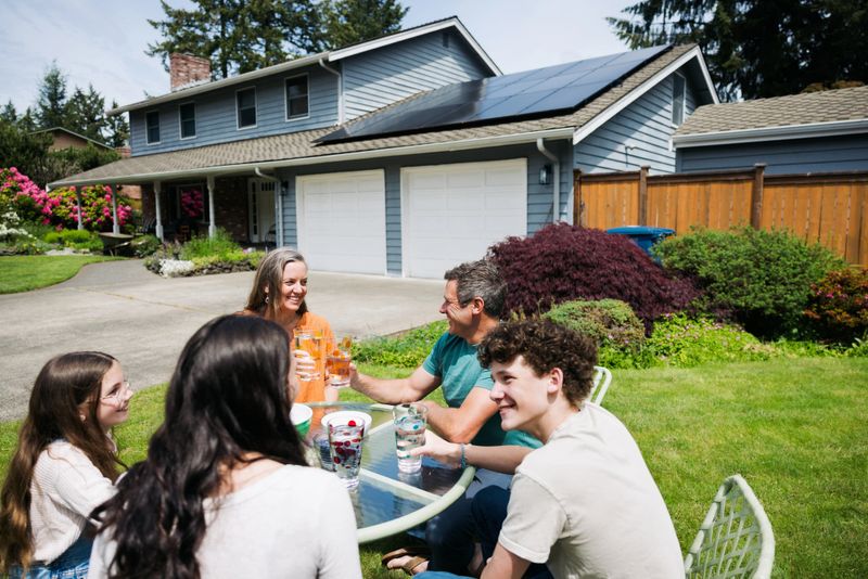 A Caucasian family with three children have fun connecting with one another outside on their lawn, sitting at an outdoor table.  Their solar panel array is visible on the roof of their home, providing green natural energy for the environmentally conscious family.  Sustainable modern lifestyle in action.