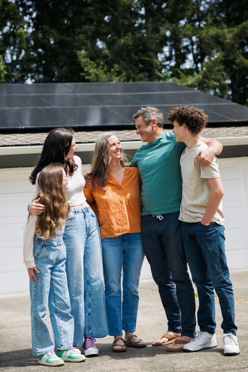 A Caucasian family with three children embrace on their driveway outside of their house.  Their solar panel array is visible on the roof of their home, providing green natural energy for the environmentally conscious family.  Sustainable modern lifestyle in action.