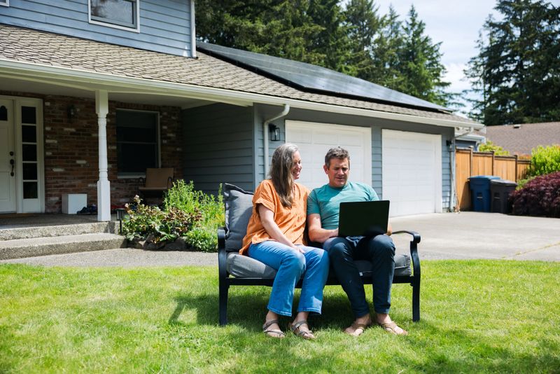 A Caucasian couple working together on a computer in their front yard on a comfortable lawn chair.  Their solar panel array is visible on the roof of their home, providing green natural energy for the environmentally conscious family.  Sustainable modern lifestyle in action.