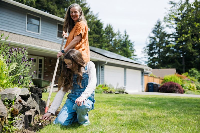 A Caucasian mother and her daughter do some weeding and pruning in their landscaped front yard.  Their solar panel array is visible on the roof of their home, providing green natural energy for the environmentally conscious family.  Sustainable modern lifestyle in action.