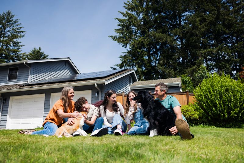 A Caucasian family with three children relax on the grass with their dogs.  Their solar panel array is visible on the roof of their home, providing green natural energy for the environmentally conscious family.  Sustainable modern lifestyle in action.
