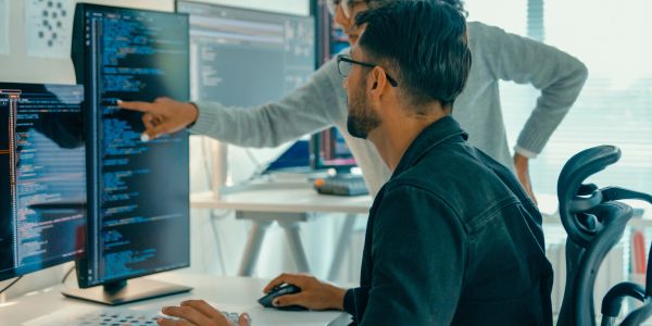 Duas pessoas analisando código em monitores duplos.