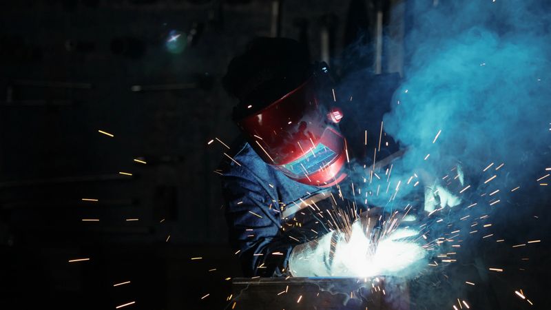 An industrial welder, clad in a safety suit and head protection, skillfully welds metal in a workshop setting. Working amidst the machinery of a car factory, sparks fly as the welder expertly maneuvers the torch. Blue-collar work in the metal industry, highlighting the precision and dedication of the worker.