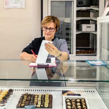 A woman behind a bakery counter writes on a clipboard, smiling.