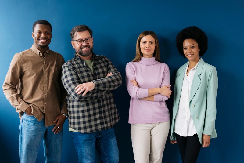 Multicultural group of people standing together and smiling against blue wall. Portrait of young men and women in casuals looking at camera.