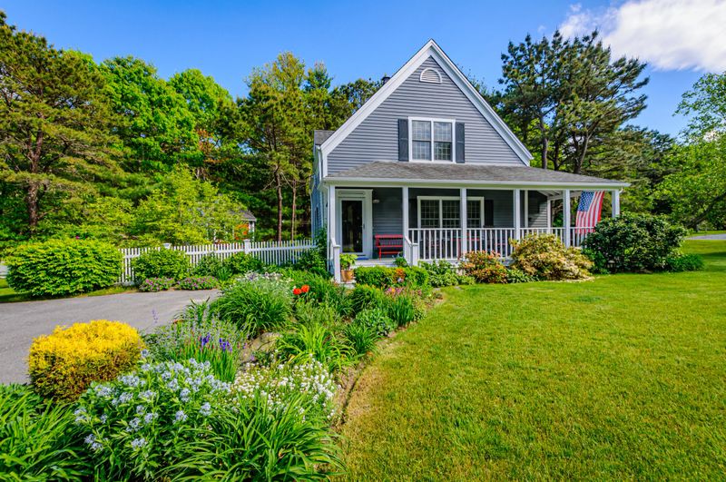 Springtime gardens along the driveway of a small single family home on Cape Cod with a flag flying from the front porch,  and the surrounding grounds,  on a late May afternoon with copy space in a clear sky area.