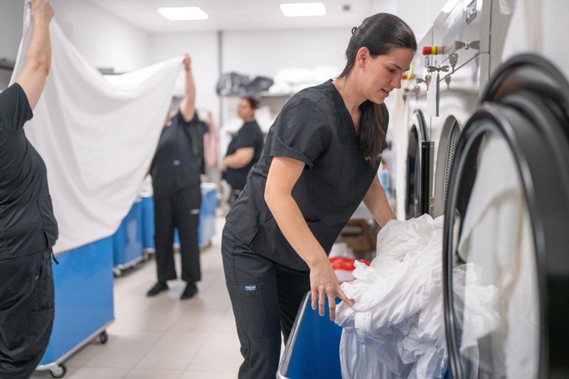 Female hotel laundry employee putting sheets into washing  machine in hotel laundry room