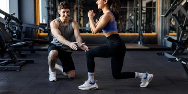 A woman doing lunges with a male trainer guiding her in a gym.
