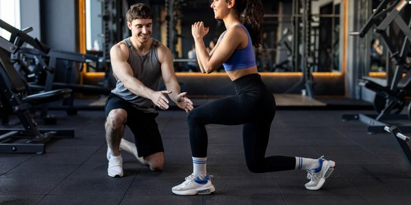 A woman doing lunges with a male trainer guiding her in a gym.