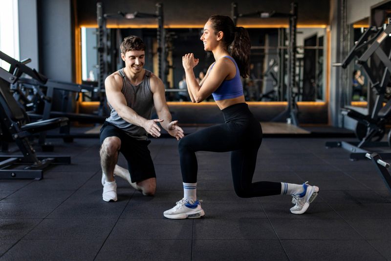 Personal trainer controlling and showing sportswoman how to do lunges correctly while kneeling in gym, side view